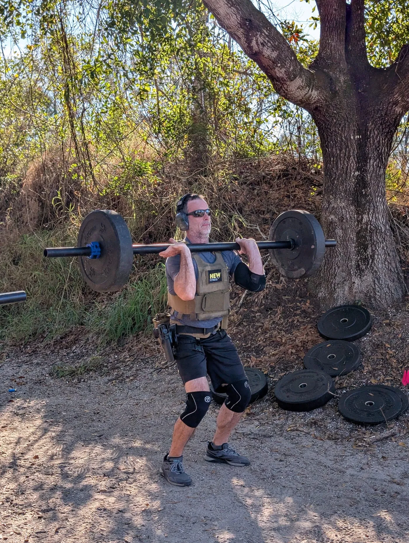 A DEFCON athlete in a plate carrier front-squatting a loaded barbell outdoors with a holstered pistol